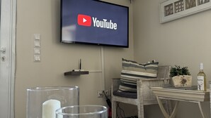 A living room with a flat-screen TV mounted on the wall displaying the YouTube logo. There are decorative glass vases with candles on a glass table in the foreground. - In the heart of Poros, stunning panoramic views, privacy and serenity. (Poros)