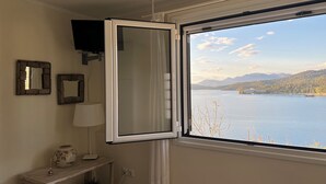 A room with a large window offering a view of a lake and mountains. There is a white shelf with a lamp and a decorative vase. - In the heart of Poros, stunning panoramic views, privacy and serenity. (Poros)