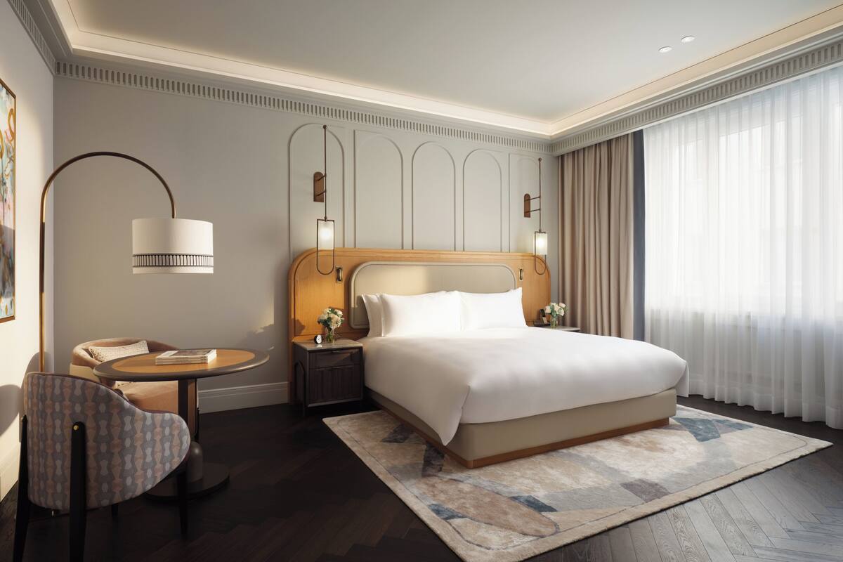 A hotel room with a large bed, a bedside table with a lamp, a chair, and a view of a building through sheer curtains.