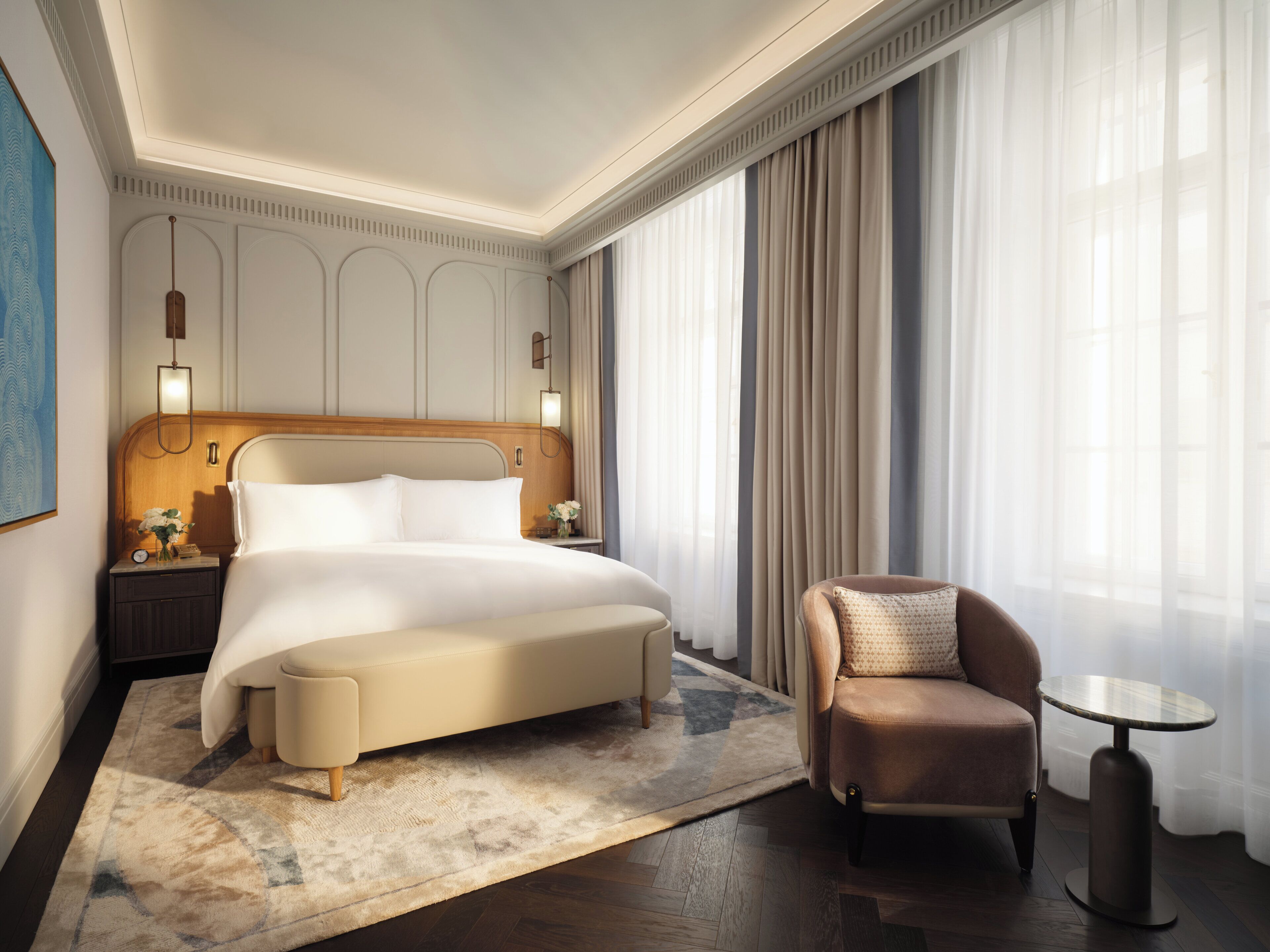 A hotel room with a large bed, a chair, a small table, and a nightstand with a lamp.