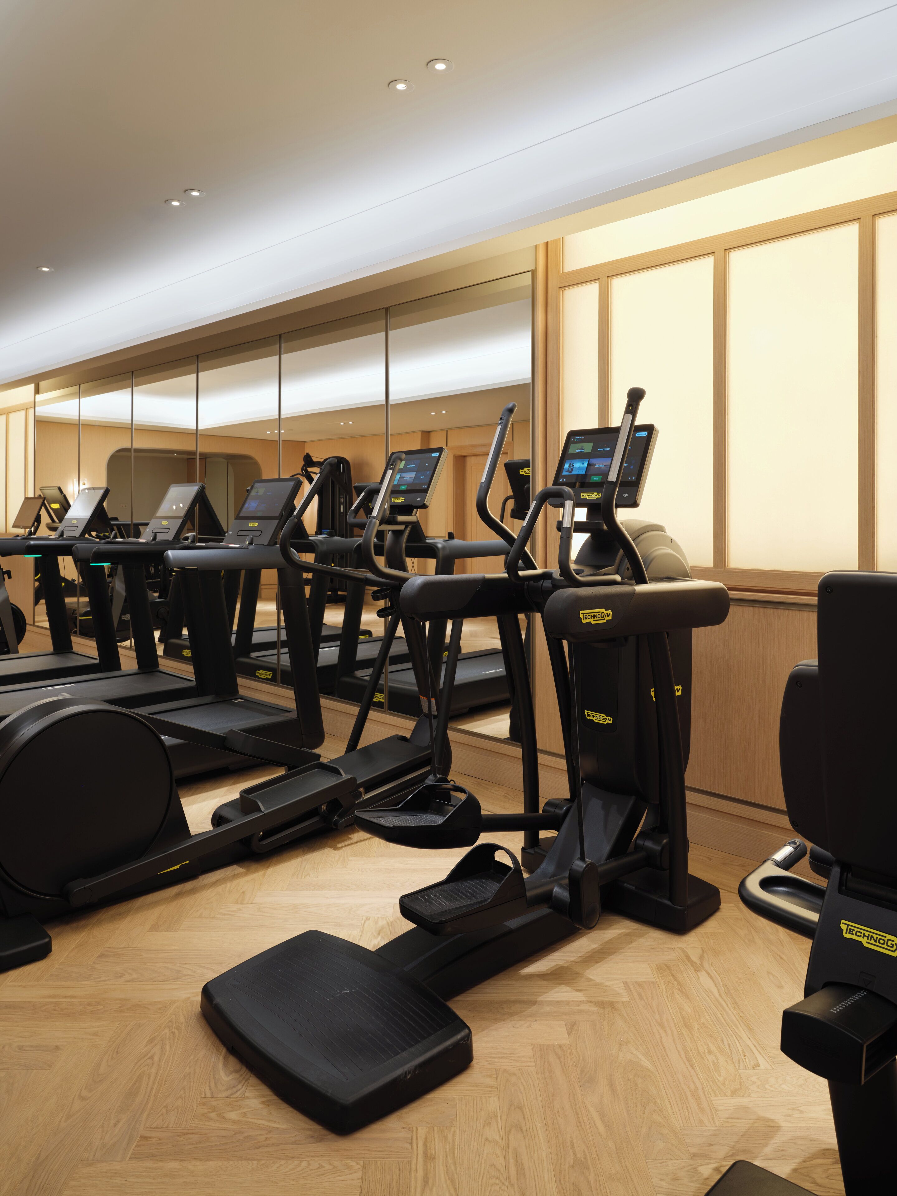 A gym with treadmills and elliptical machines, wooden floors, and a large mirror wall.