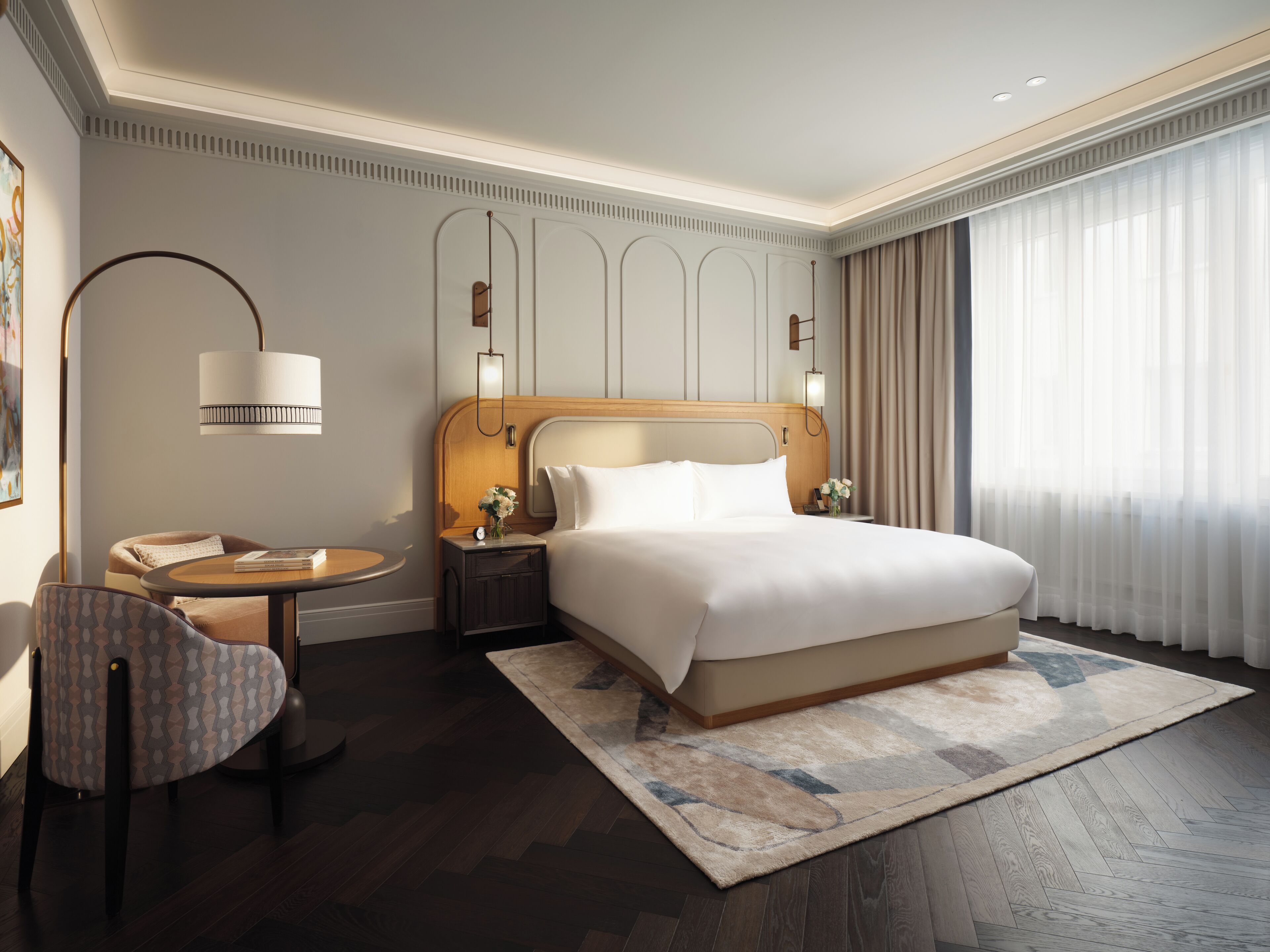 Quarto de hotel com uma cama grande, mesa de cabeceira com luminária, uma cadeira e vista para o exterior através de cortinas translúcidas.