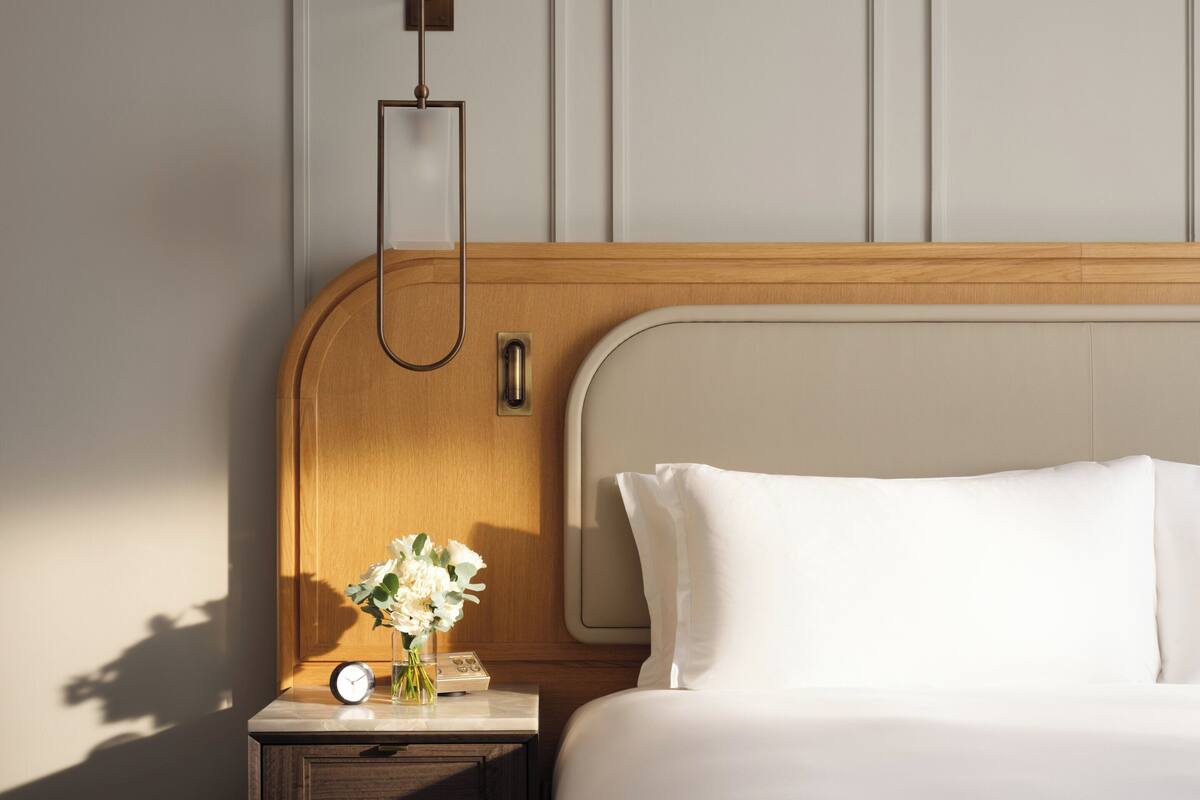 A hotel room with a wooden headboard, a bedside table with a clock and a vase of flowers, and a bed with white bedding.