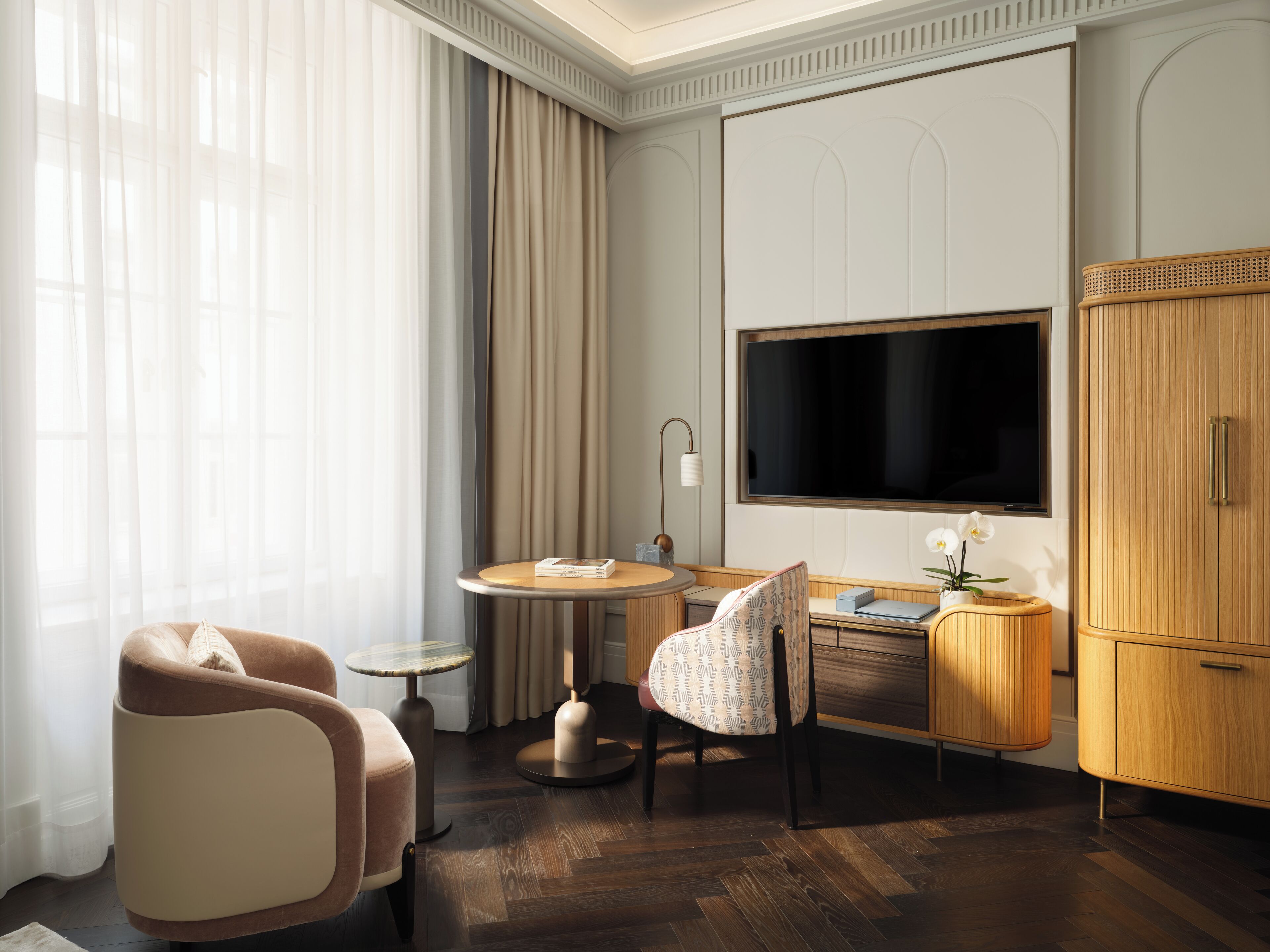 Una habitación de hotel moderna con un televisor de pantalla plana grande, una mesa redonda de madera, un sillón cómodo, una mesita redonda pequeña y un mueble de madera.