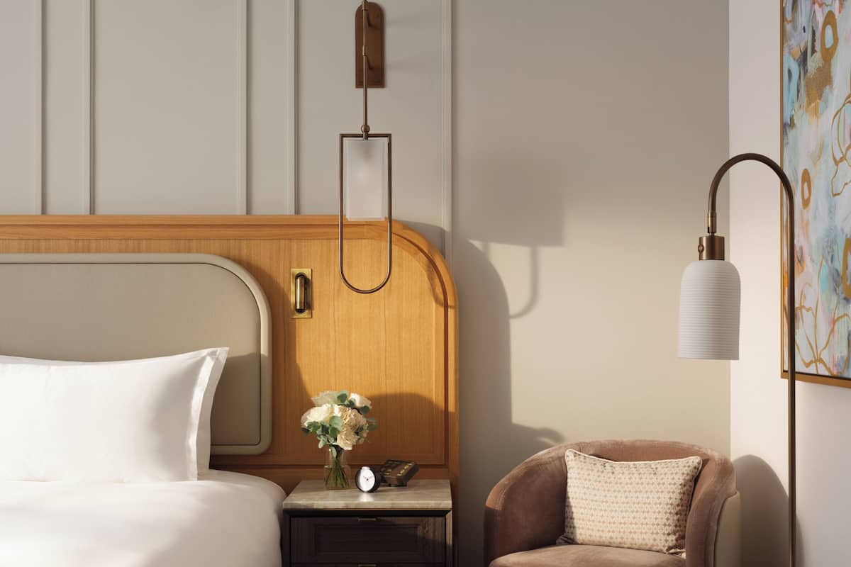 Ein Hotelzimmer mit hölzernem Kopfteil, einem Bett mit weißer Bettwäsche, einem Nachttisch mit Blumenvase, einem braunen Leder-Sessel, einem runden Tisch und einer modernen Stehlampe.