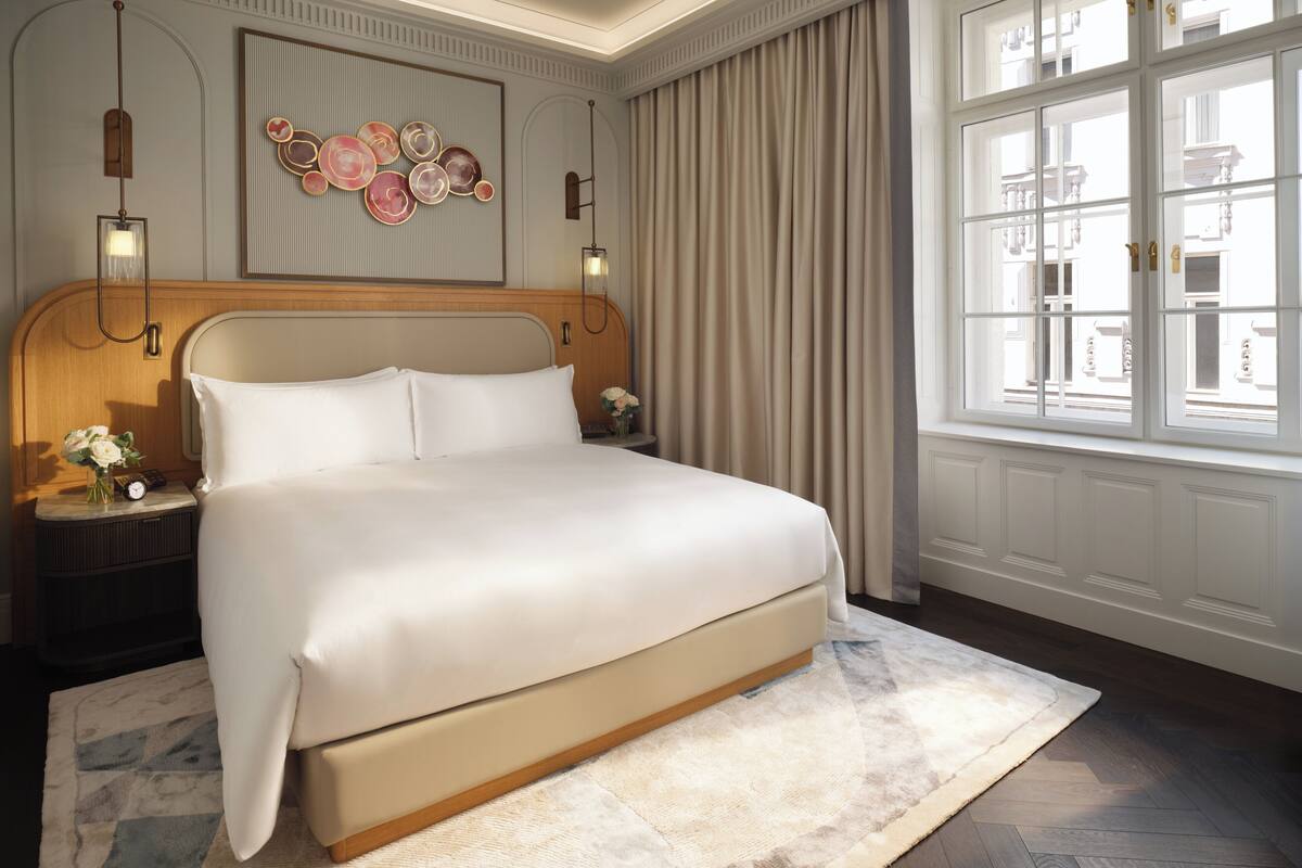 A hotel room with a large bed, a nightstand with a clock, a vase of flowers, and a window with curtains.