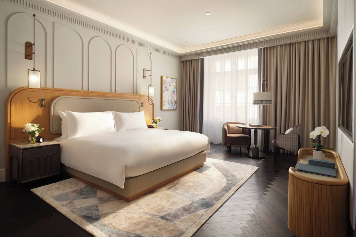 A modern hotel room with a large bed, a bedside table with a lamp, a small round table, and a chair.