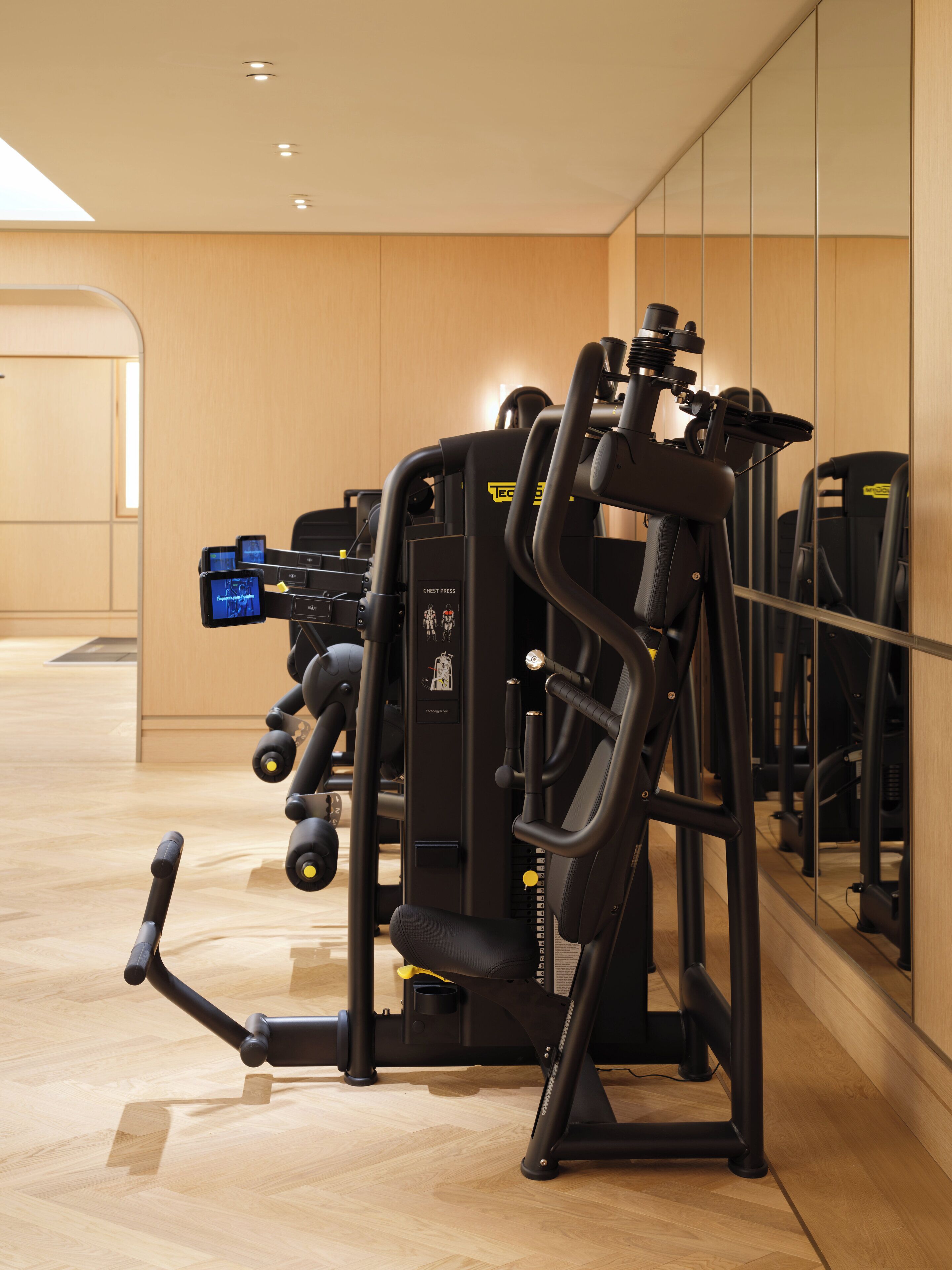 Uma academia com diversos equipamentos de exercício, uma tela digital e piso de madeira.