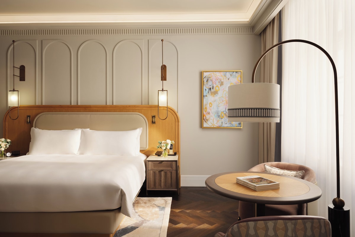 Une chambre d’hôtel avec un grand lit, des tables de chevet, une table ronde avec une chaise et une lampe sur pied.