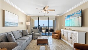Un salon comprenant un canapé, un pouf, des fauteuils, un ventilateur de plafond, un téléviseur et une vue sur l’océan grâce à de grandes fenêtres.