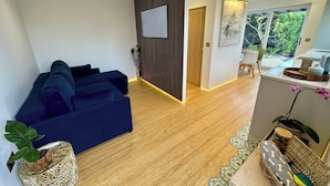 Un salón moderno con un sofá azul, suelo de madera, televisor montado en la pared y comedor con vistas a zonas verdes.