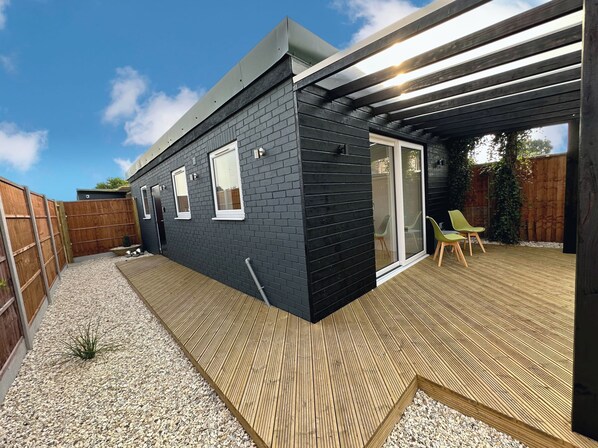 Een modern huis met een zwarte gevel, een houten terras en een schuifdeur naar een zitgedeelte buiten.