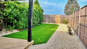 Un patio trasero bien cuidado con una terraza de madera, césped verde y un camino de grava.