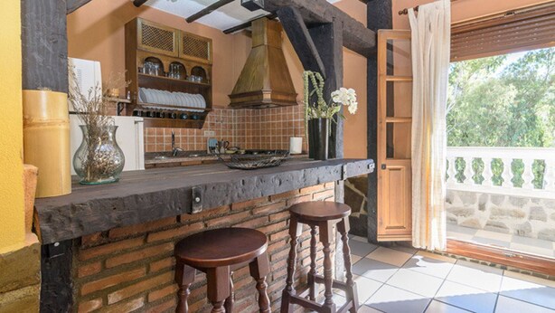 A kitchen with a brick wall, wooden stools, a hanging pot rack, and a view of greenery outside.
