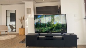 A modern living room with a large flat-screen TV on a black entertainment unit, a wooden floor, a beige sofa, and a decorative mirror.