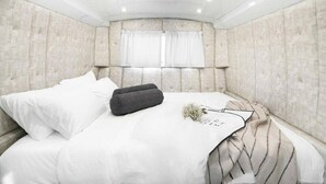 A modern bedroom with a tufted headboard, a bed with white bedding, a dark throw pillow, and a decorative item on the bed. - The Daydream Bus Hideaway by Sea N' Rent (Gesher HaZiv)