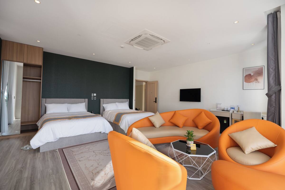 Una camera d'albergo moderna con due letti, un divano arancione, un tavolino e una TV a parete.