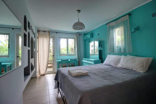 Un dormitorio con cama, escritorio y silla. Hay ventanas con cortinas, una lámpara en el techo y vistas a zonas verdes al exterior.