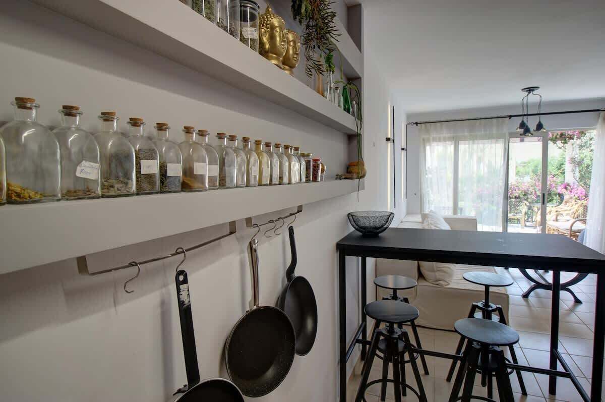 Una cocina con estantes llenos de frascos de vidrio, una mesa negra con taburetes y ollas colgando.