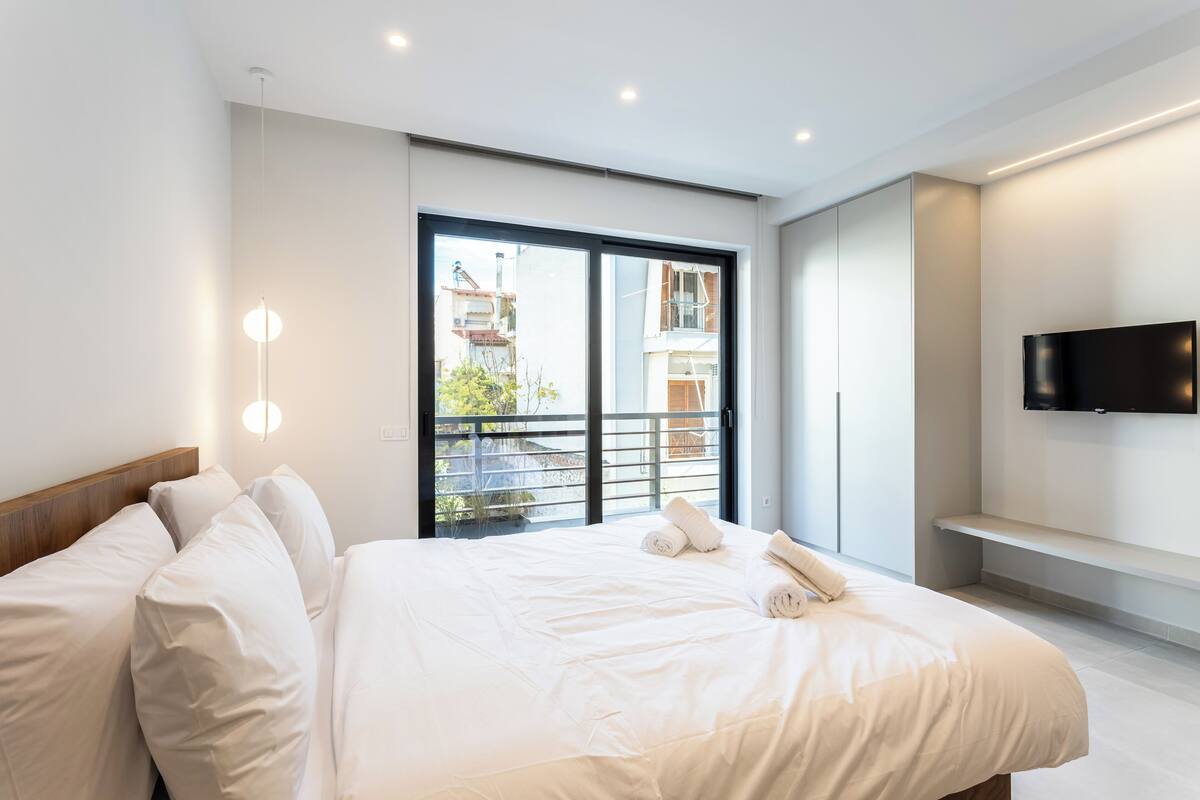 Una habitación de hotel moderna con una cama grande, un televisor montado en la pared y un balcón con vistas a la ciudad.