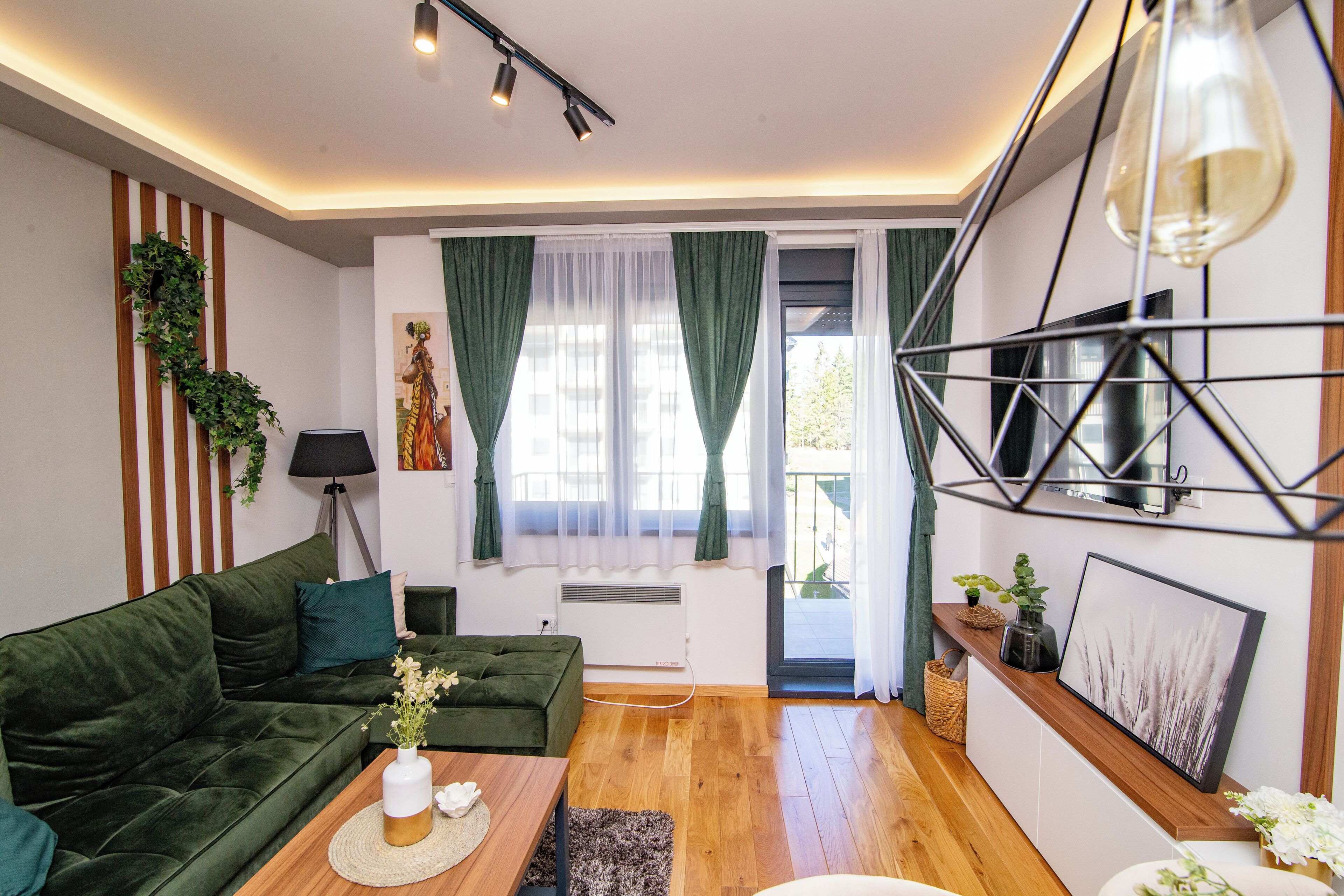 A modern living room with a green sofa, a coffee table, and a TV. There is a window with curtains, a plant, and a picture frame on the wall.