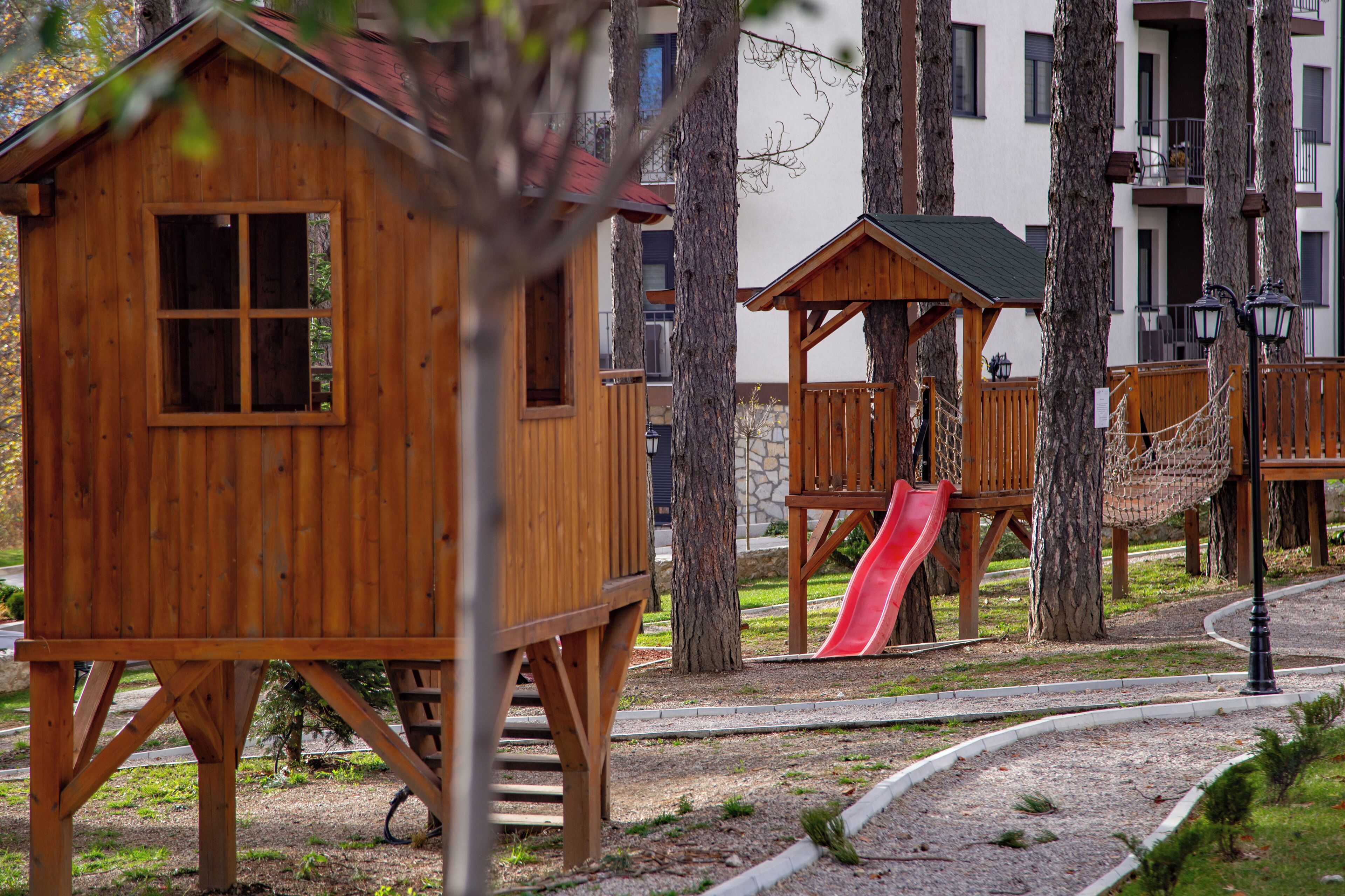 A wooden playhouse with a window, a red slide, and a rope bridge in a park area with trees and a building in the background.