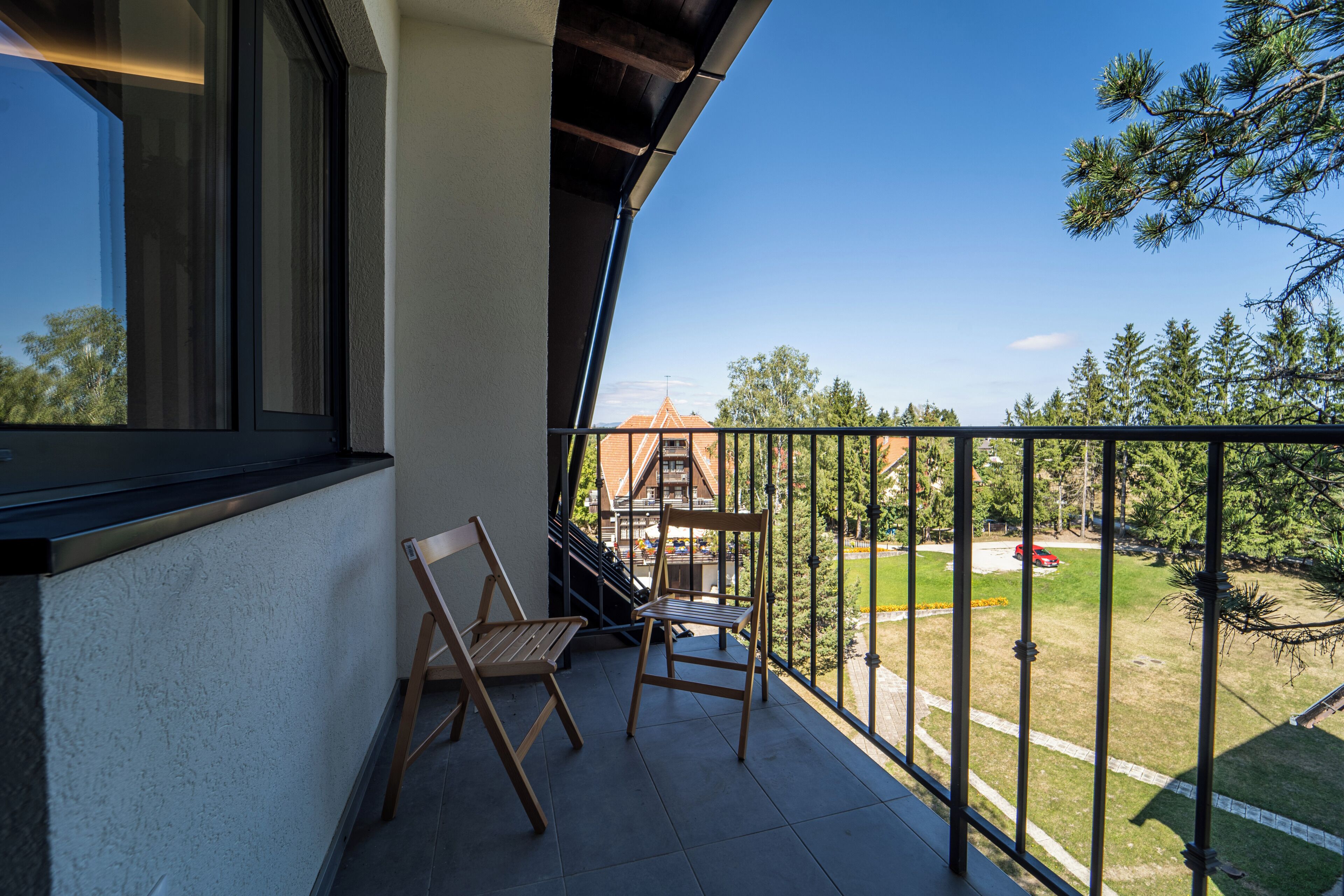 A balcony with two wooden chairs, a view of a building with a red roof, and a clear blue sky.
