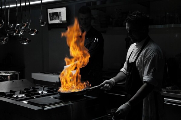 Un cocinero preparando comida en una sartén sobre la estufa, con llamas visibles, en una cocina.