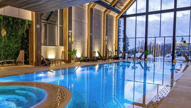 A modern indoor swimming pool with clear blue water, large windows offering views of the outdoors, and lounge chairs along the poolside.