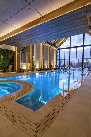 Una piscina interior moderna con agua azul cristalina, grandes ventanales que ofrecen vistas al exterior y sillones de descanso a orillas de la piscina.