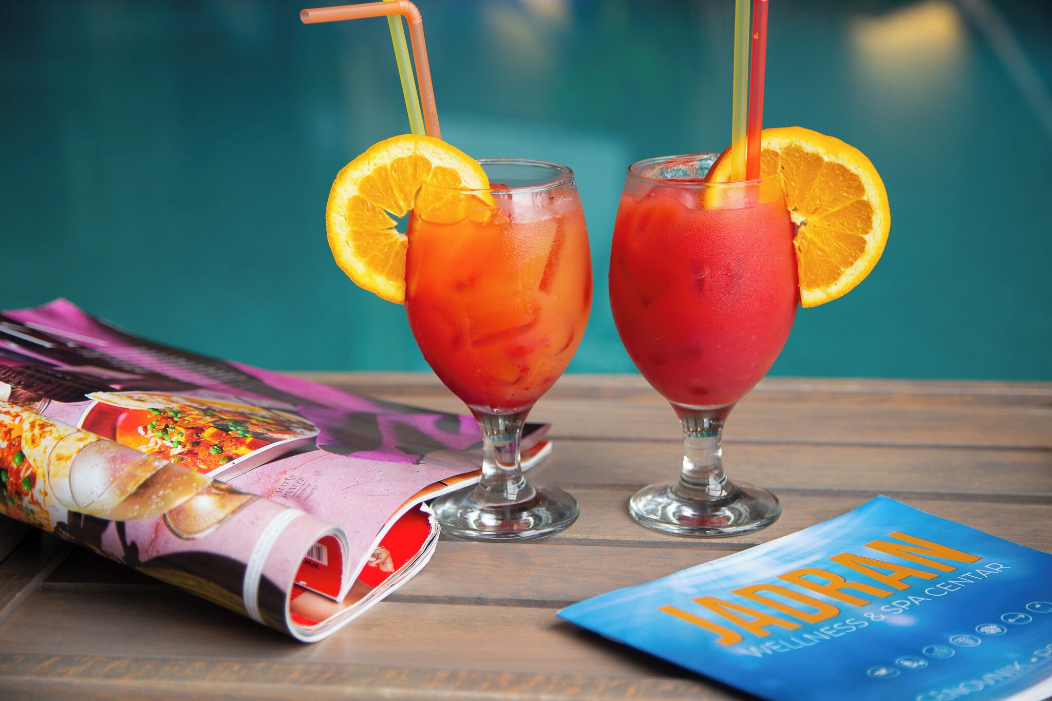 Two drinks with orange slices and straws, a magazine, and a brochure on a wooden table.