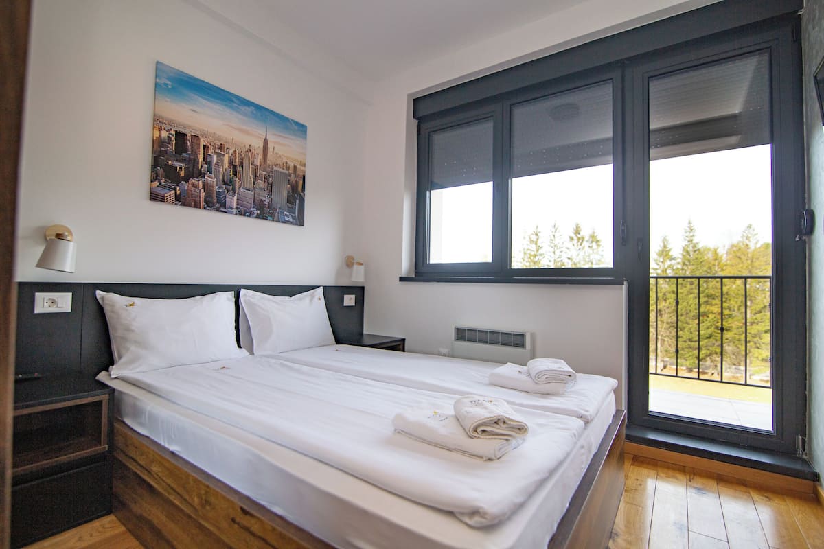 A double bed with white linens, a nightstand with a lamp, a painting of a cityscape, and a window with a view of trees.