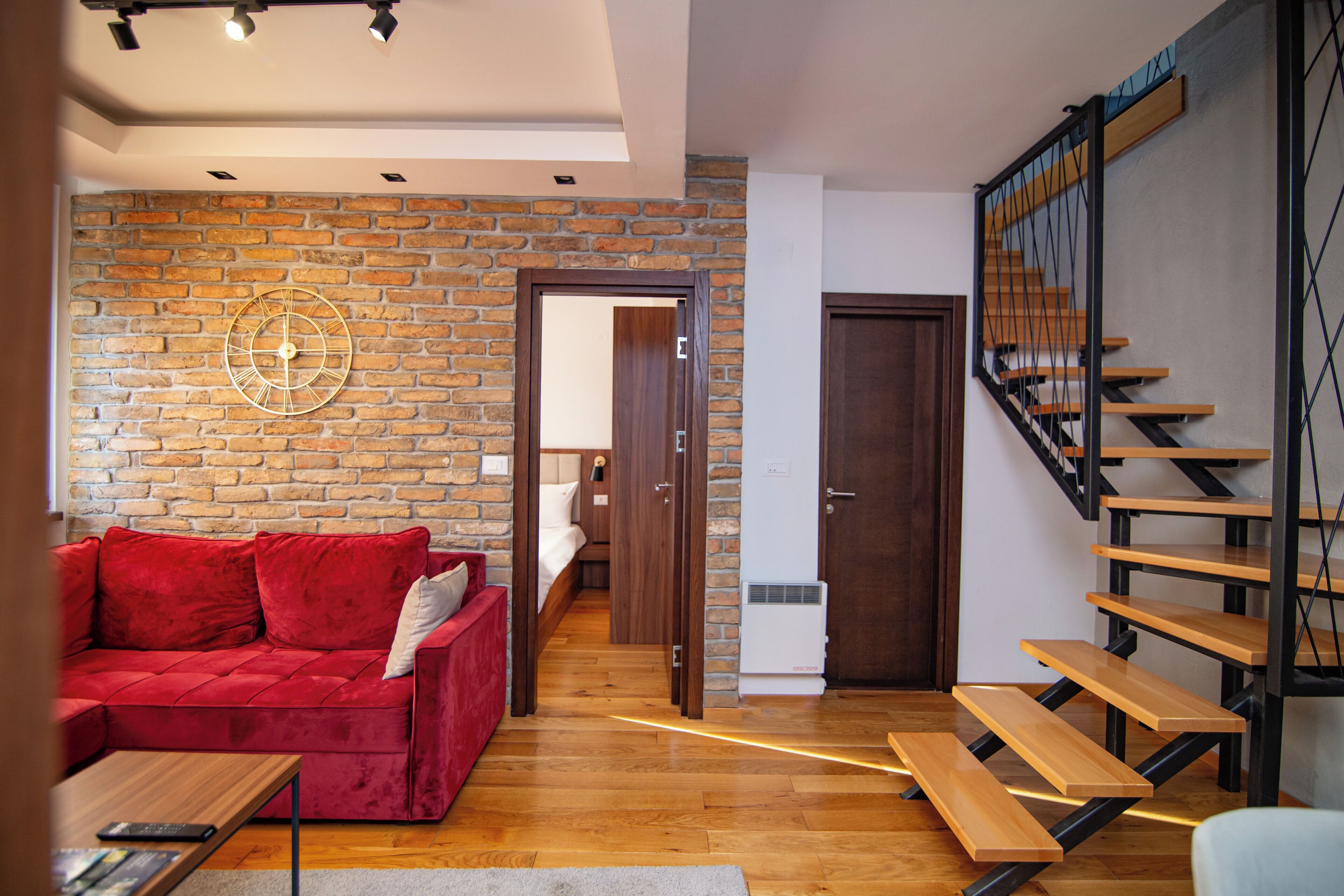 A modern living room with a red sofa, wooden flooring, a brick wall, and a staircase with wooden steps and metal railings.