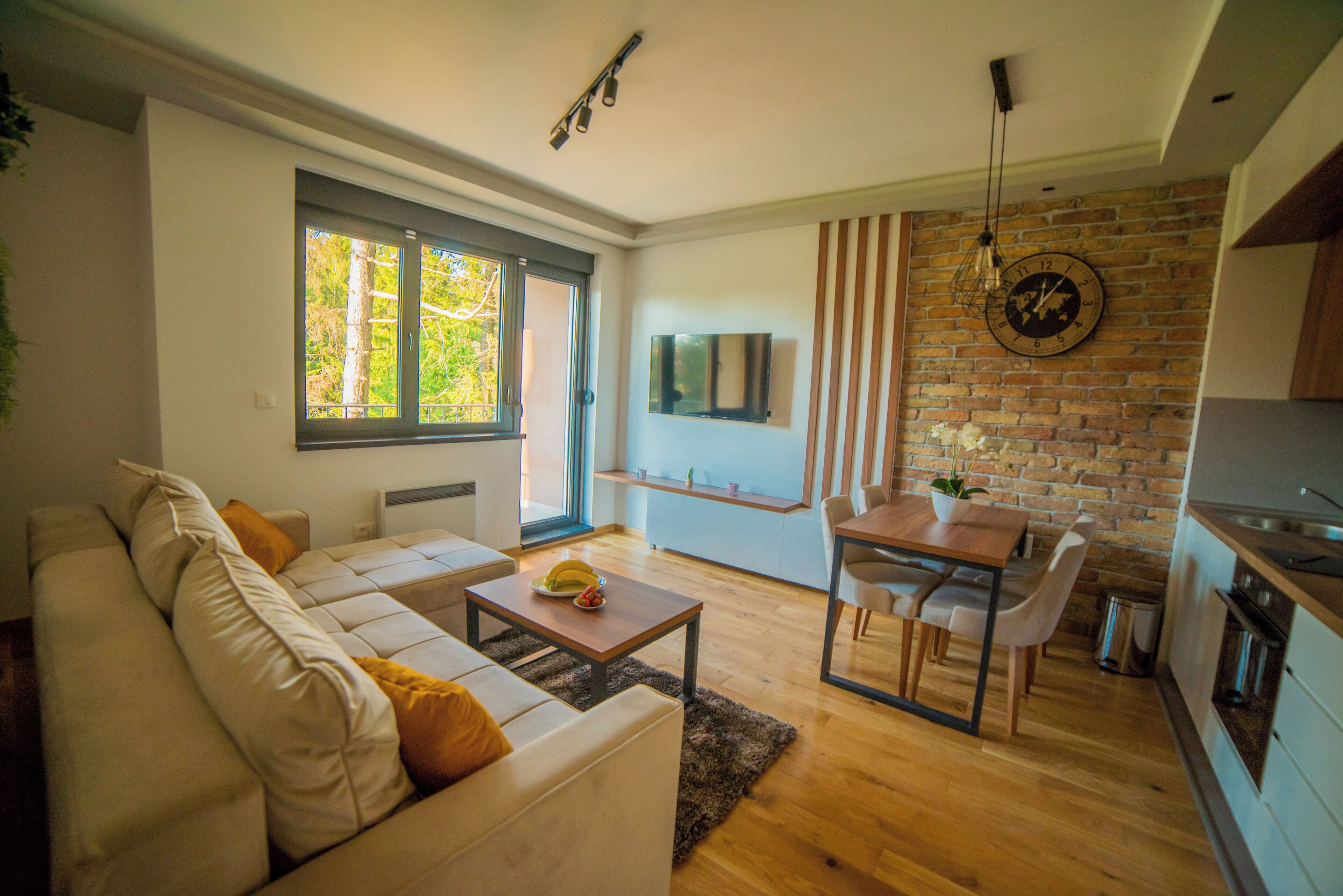A modern living room with a sofa, coffee table, and a dining area with a table and chairs. There is a TV mounted on the wall and a brick wall in the background.