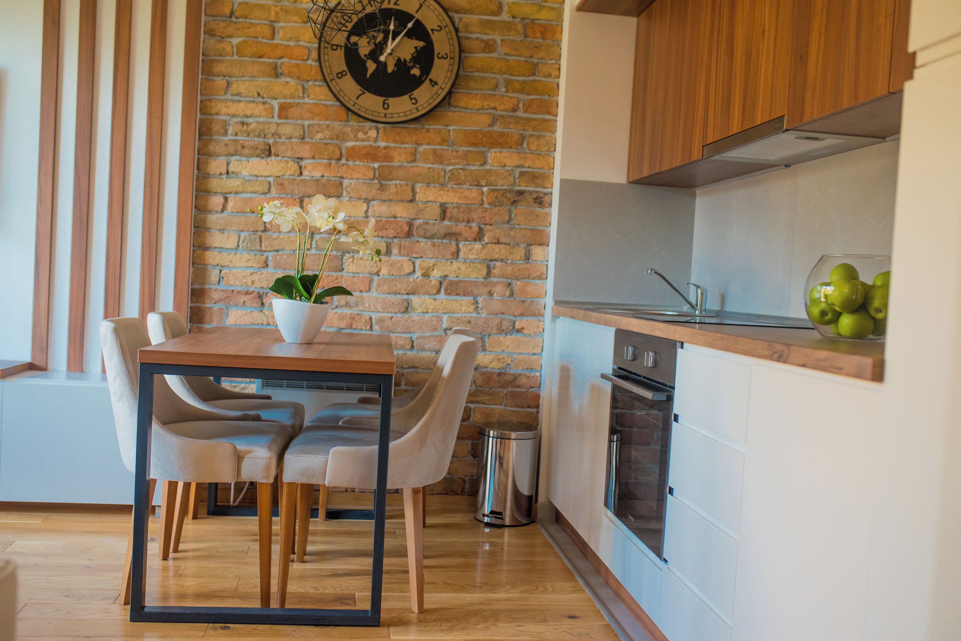 A modern kitchen with a brick wall, wooden cabinets, a dining table with chairs, and a clock.