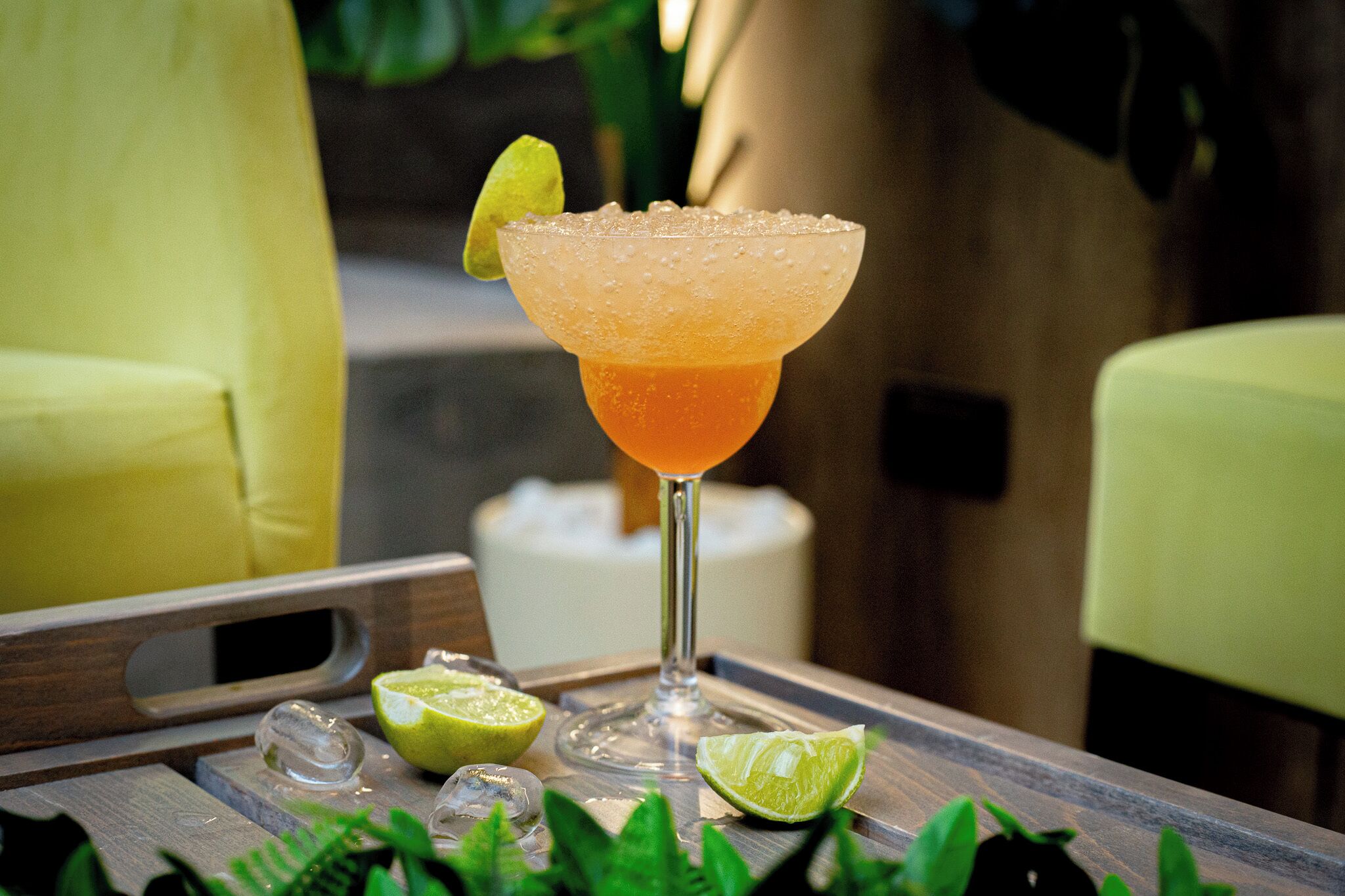 A cocktail glass with a lime garnish on a wooden tray with ice cubes and lime halves.