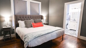 Ein Schlafzimmer mit einem gesteppten Kopfteil, einem Bett mit weißer Bettwäsche, grau-roten Kissen, einer gemusterten Tagesdecke und einem Nachttisch mit einer Lampe.