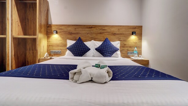 A neatly made bed with a blue and white bedspread, two blue pillows, and a towel with a green tag. There are two bedside tables with a water bottle and a lamp on each. - WIND FLOWER (Mumbai)