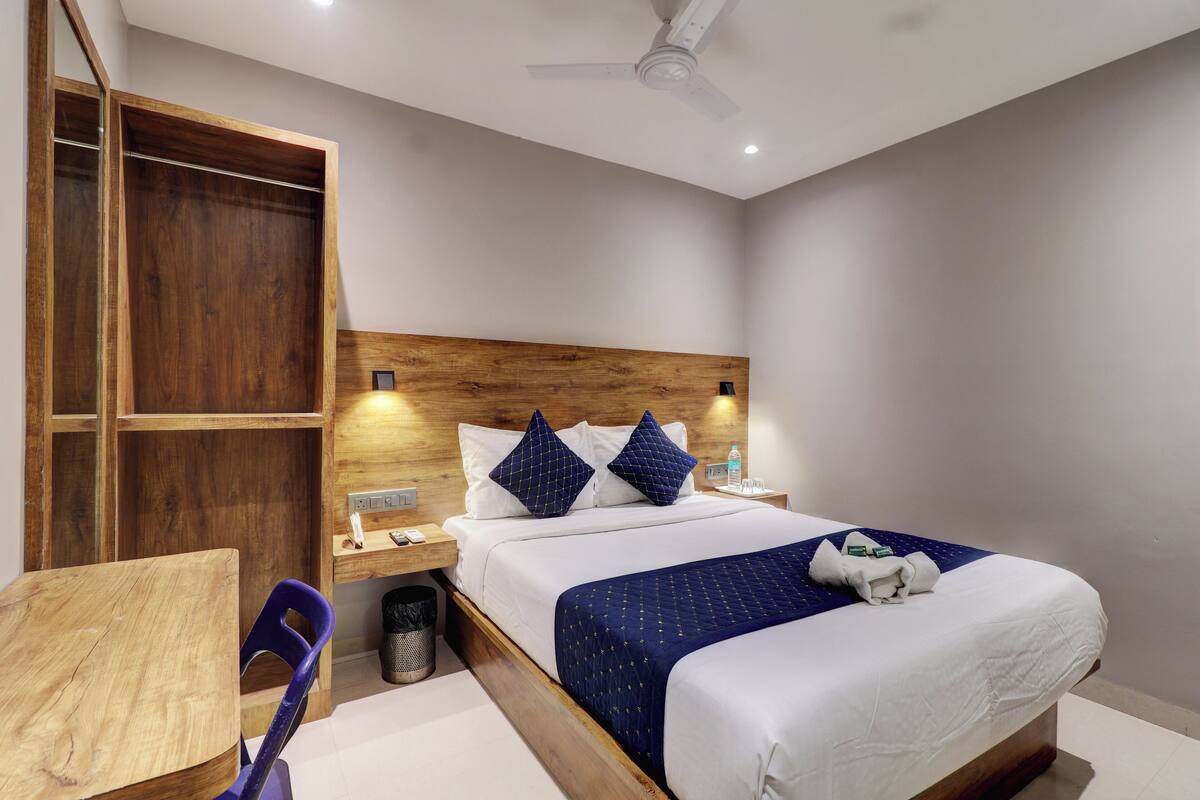 A hotel room with a wooden headboard, a bed with white and blue bedding, a small desk, and a chair.