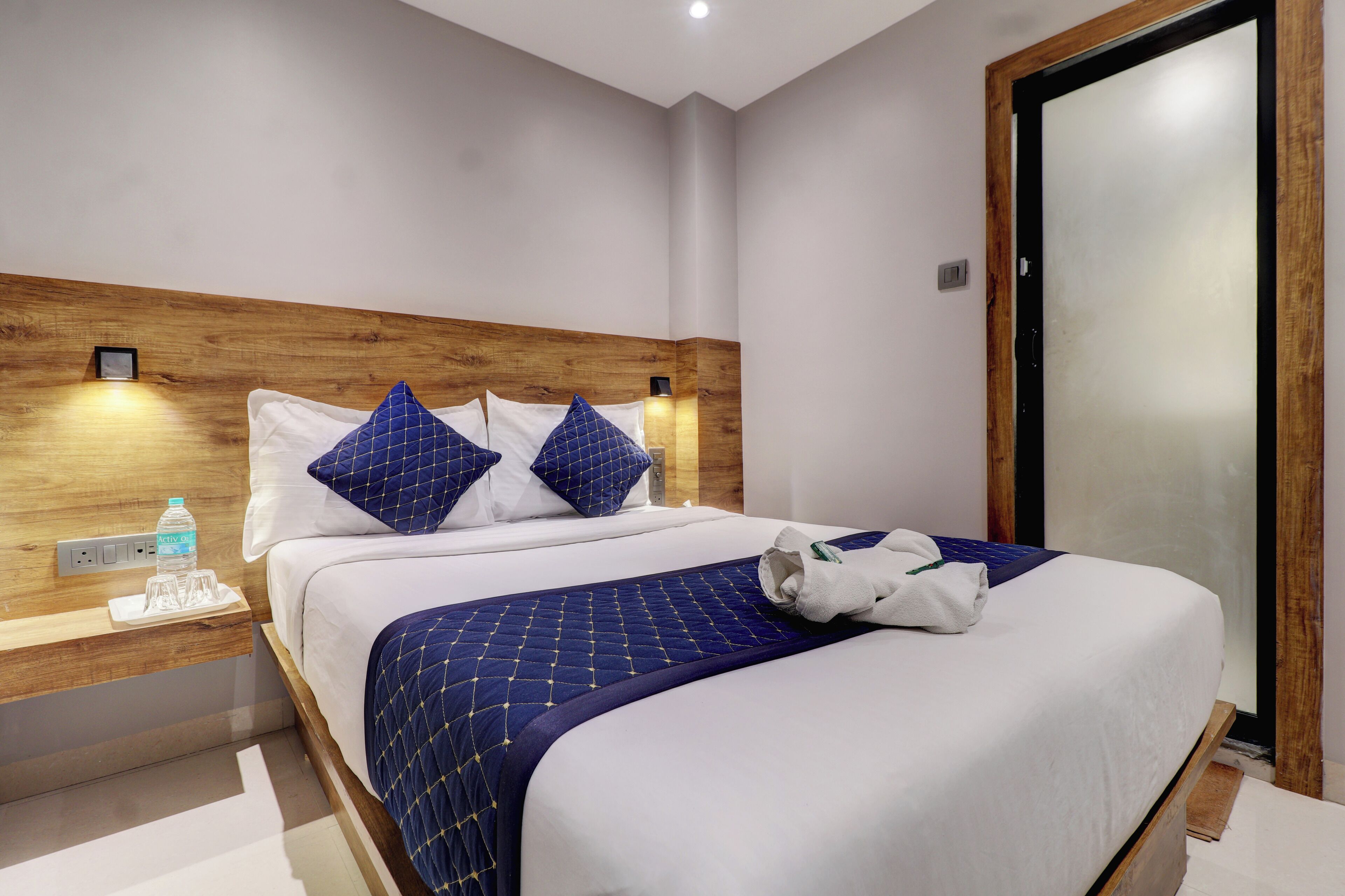 A hotel room with a wooden headboard, a bed with white and blue bedding, a water bottle on the nightstand, and a towel on the bed.