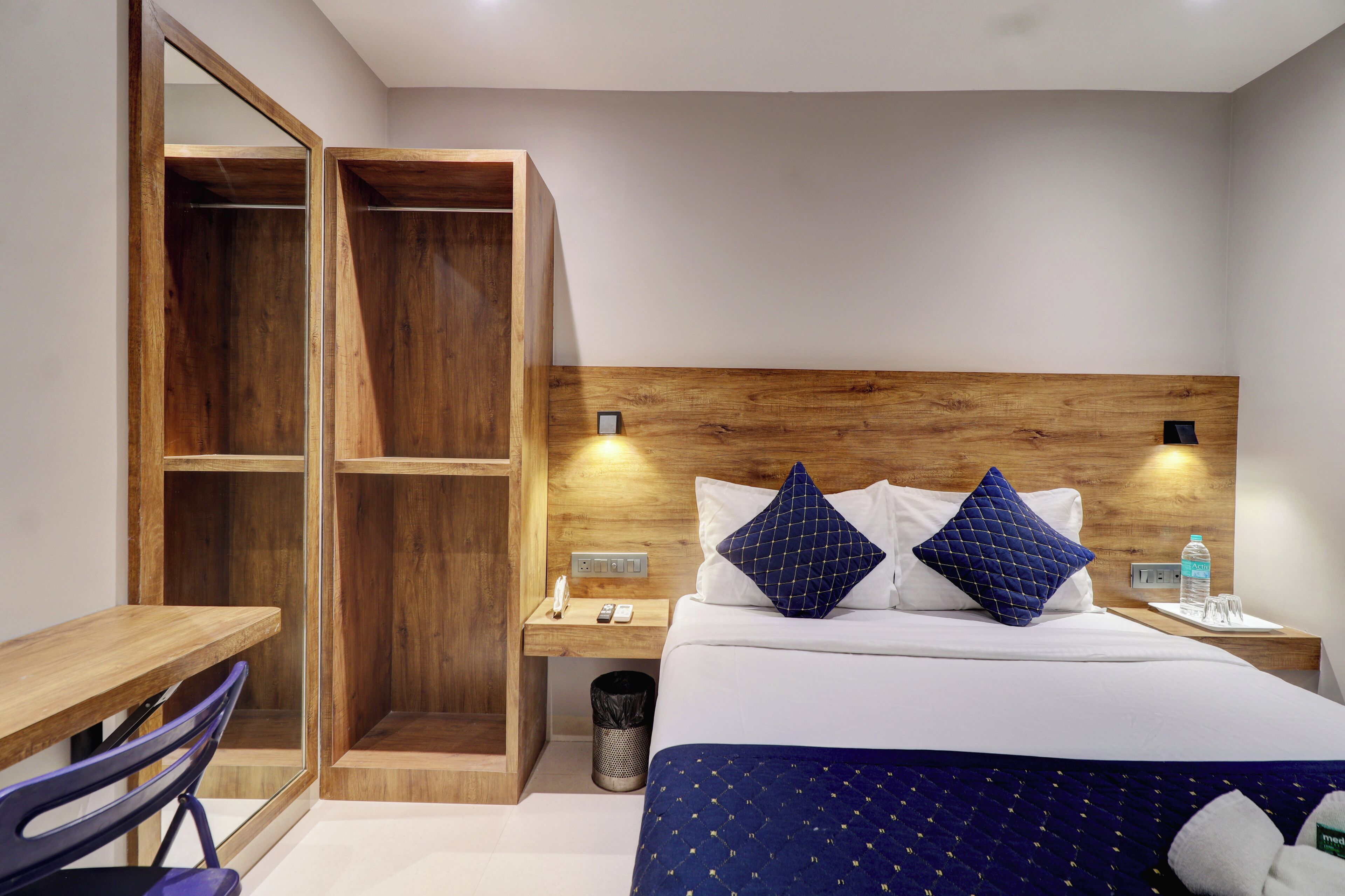 A hotel room with a wooden headboard, a bed with two pillows, a bedside table with a lamp, a small trash bin, and a desk with a chair.