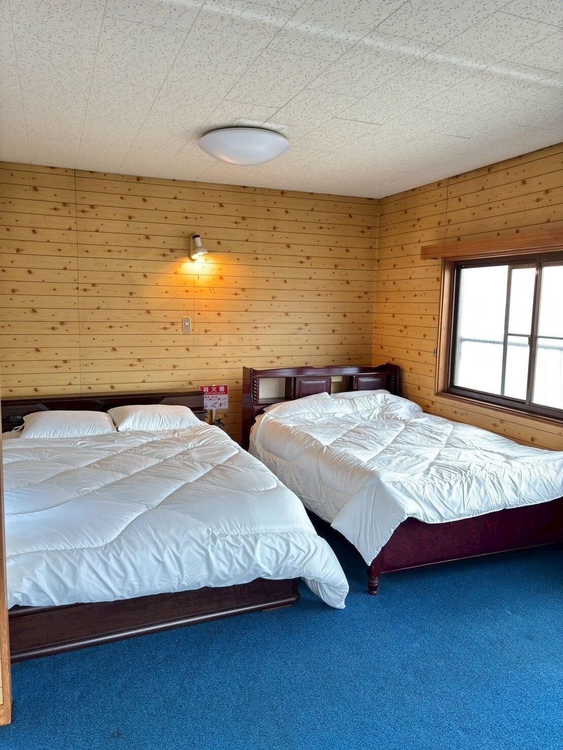 Une chambre avec deux lits, des murs en bois, un luminaire au plafond et une fenêtre.