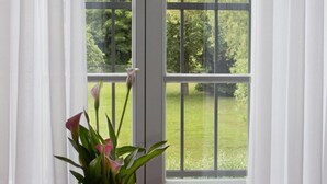 A window with white curtains and a view of a green lawn and trees. - Hotel Gutshof (Herborn)