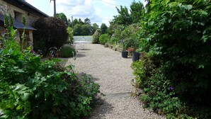 Un chemin de gravier mène à une clôture blanche, entourée d’une végétation luxuriante et de plantes en pot.