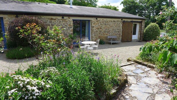 Un bâtiment en pierre avec une allée de gravier, une table et des chaises en métal blanc, et une variété de plantes à fleurs.