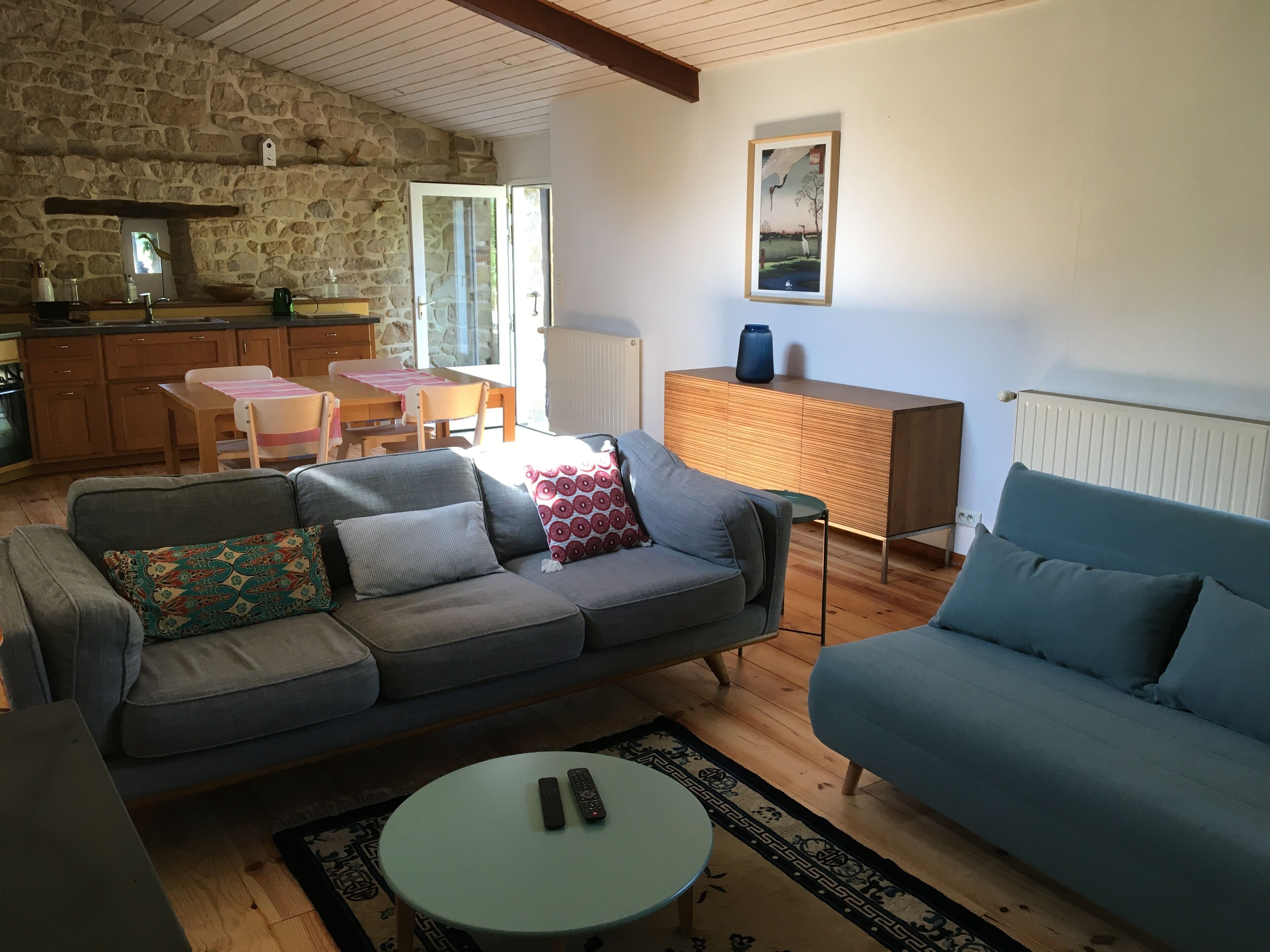 A living room with a grey sofa, a blue sofa, a small round table, and a kitchen area with wooden cabinets and a stone wall.