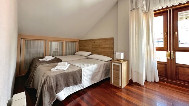 Un dormitorio con cabecera de madera, cama con duvet, mesita de noche con lámpara y ventana con cortinas.