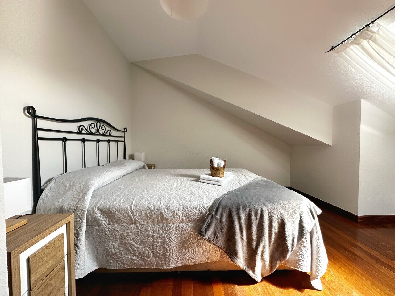 A bedroom with a sloped ceiling, a bed with a patterned bedspread, a bedside table, and a small basket with towels.