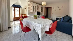 Un comedor con una mesa cubierta por un mantel estampado, rodeado de sillas rojas. Se observa un sofá azul y una zona de cocina al fondo.