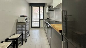 Cocina estrecha con microondas, horno y electrodomésticos de acero inoxidable. Hay una encimera de madera y una puerta corredera que da a un balcón.